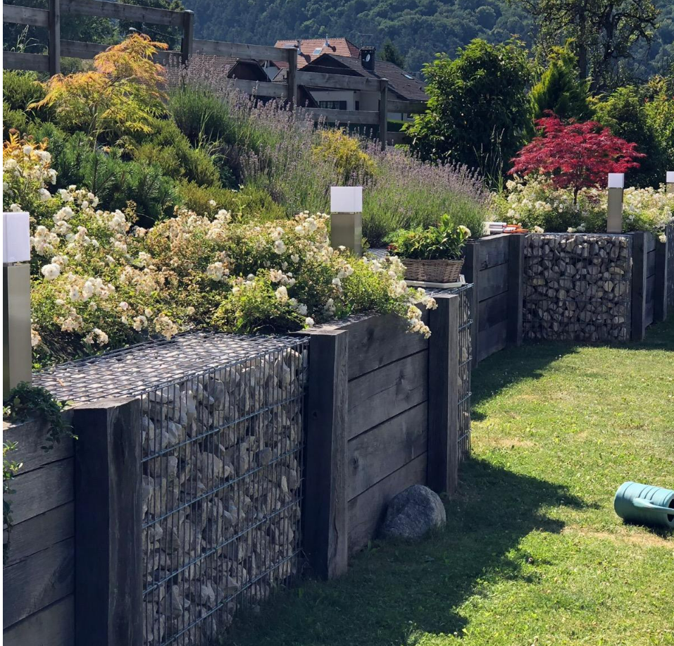 Un mur de soutènement à plusieurs niveaux, composé de gabions en fil de fer remplis de pierres et de poteaux en bois sombre, est installé dans un jardin paysager ensoleillé.