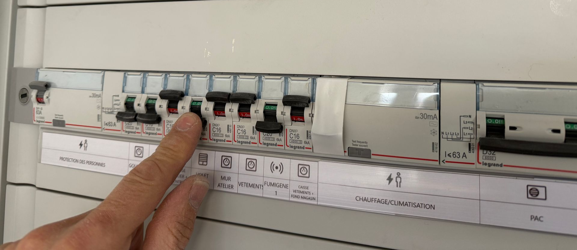 Le doigt d'une personne désigne l'un des nombreux interrupteurs noirs d'un panneau de disjoncteurs électrique blanc.