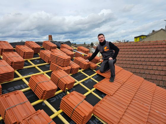 Toit de tuiles brunes d'une maison en construction, vu de la crête, avec les maisons du quartier en arrière-plan.