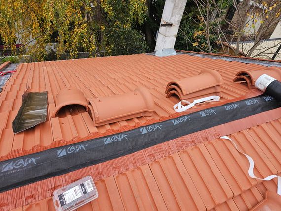 Toit en pente recouvert de tuiles, avec un puits de lumière et une échelle, sous un ciel ensoleillé.