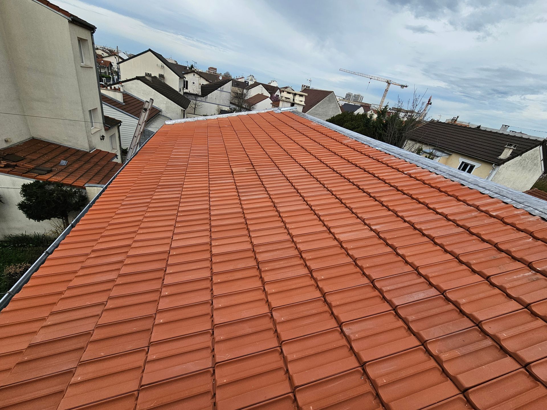 Un ouvrier portant un casque de chantier jaune est agenouillé sur un toit sous un ciel bleu nuageux, utilisant un outil.