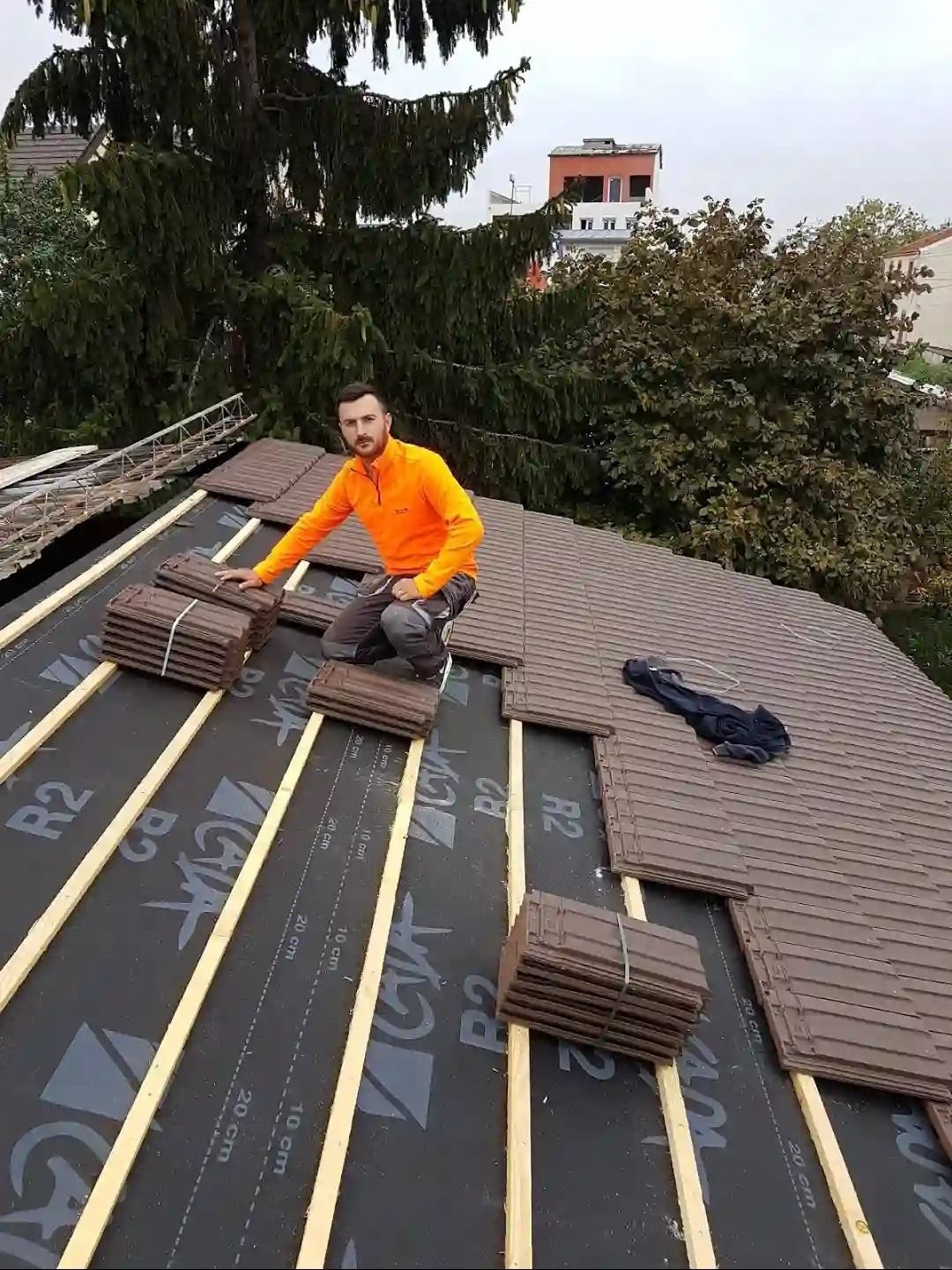 Un ouvrier en veste orange pose du feutre bitumé sur un toit pentu, avec des arbres et une maison en arrière-plan.