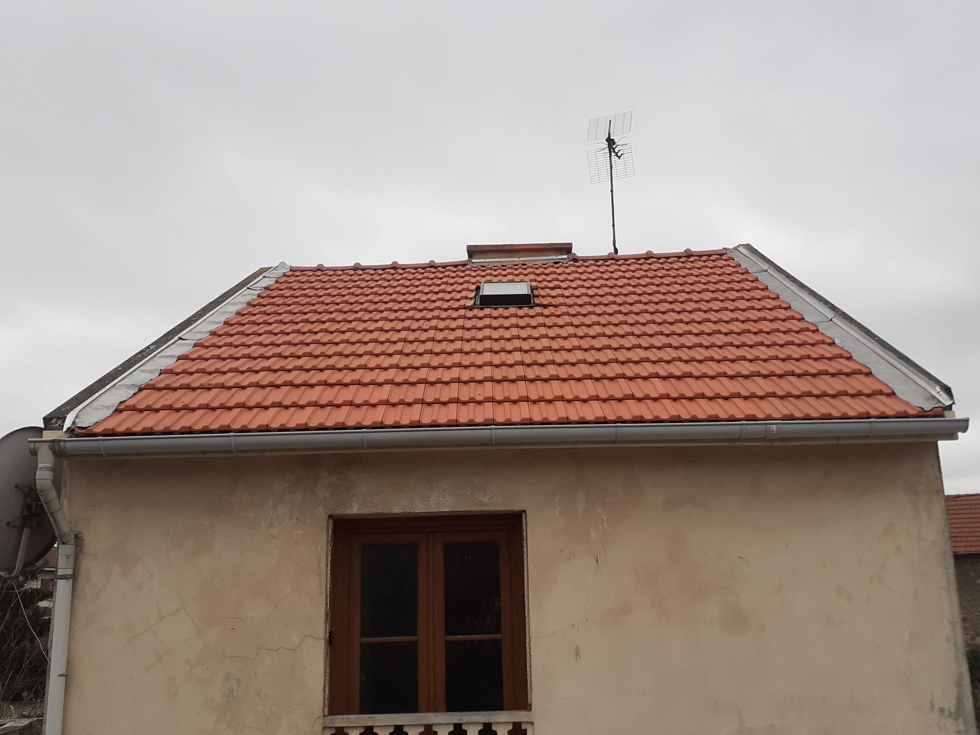 Petite maison beige au toit de tuiles rouges, avec une fenêtre et une antenne se détachant sur un ciel gris.