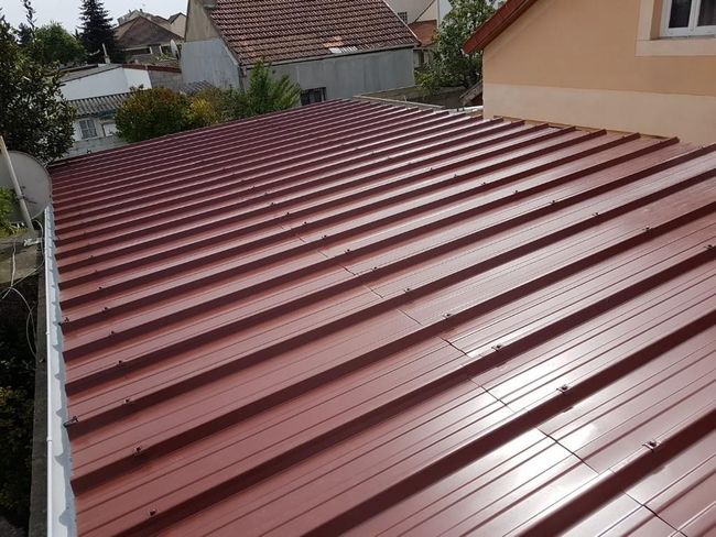 Toiture en tôle rouge d'une maison, vue du ciel, avec des arbres et des bâtiments en arrière-plan.