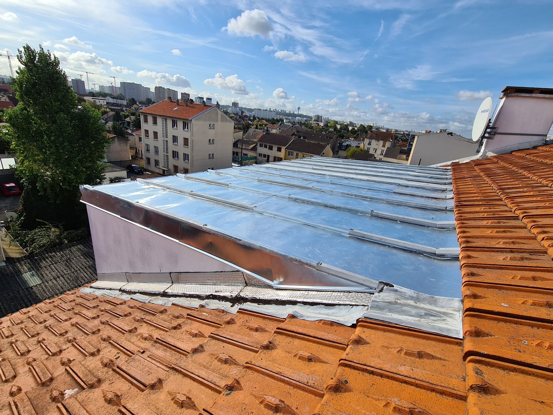 Vue depuis un toit avec des tuiles orange et des plaques métalliques et des bâtiments de la ville au loin sous un ciel bleu.
