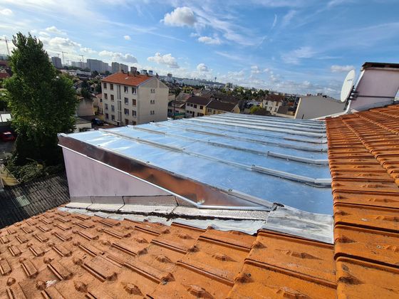 Vue depuis un toit aux tuiles orange, avec des bandes métalliques et des immeubles sous un ciel bleu.