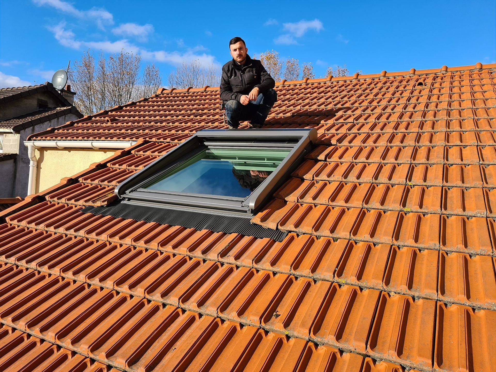 Un homme inspecte une fenêtre sur un toit de tuiles rouges par une journée ensoleillée.