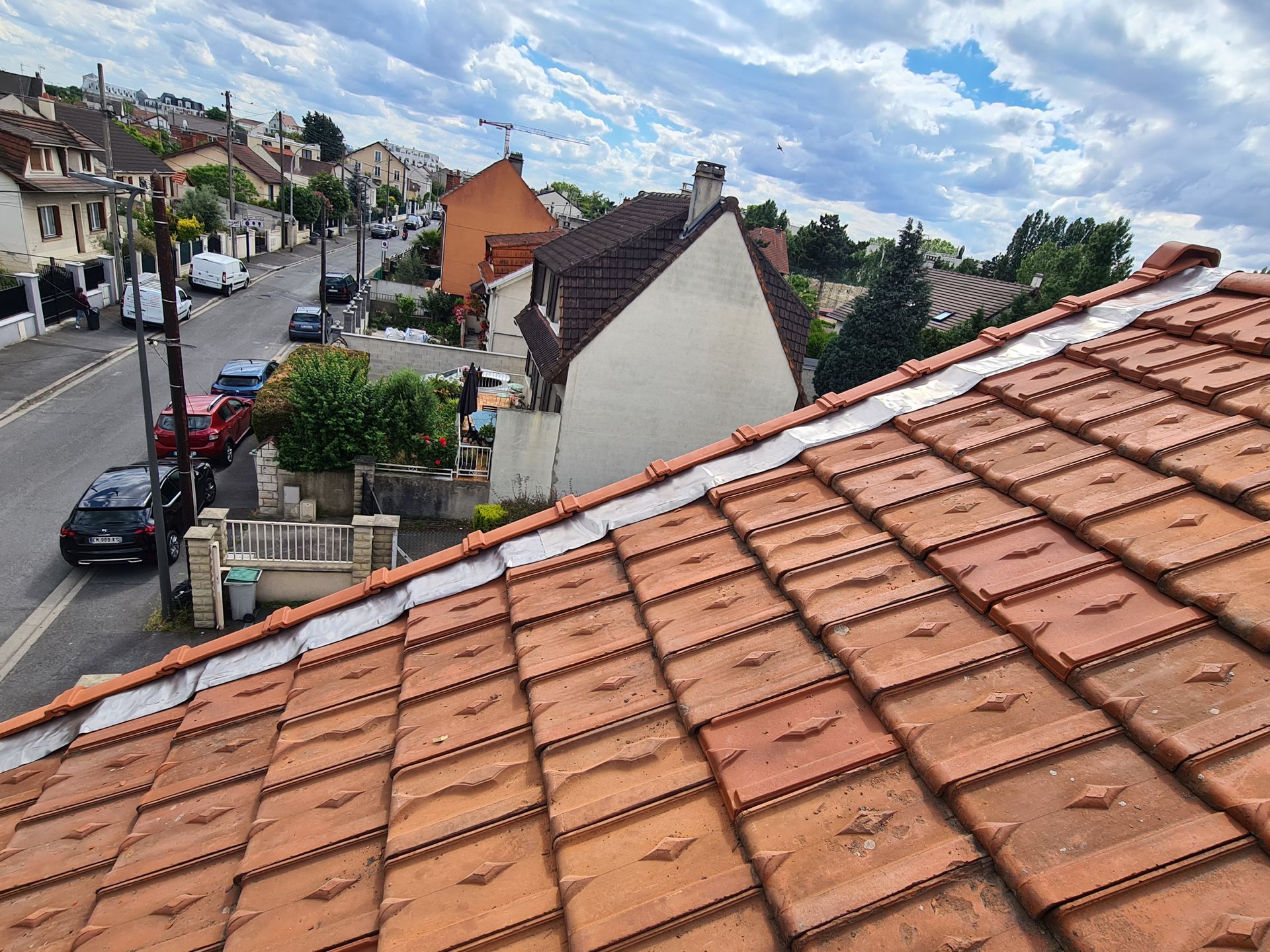 Vue depuis un toit de tuiles rouges sur une rue résidentielle bordée de maisons et de voitures garées sous un ciel nuageux.
