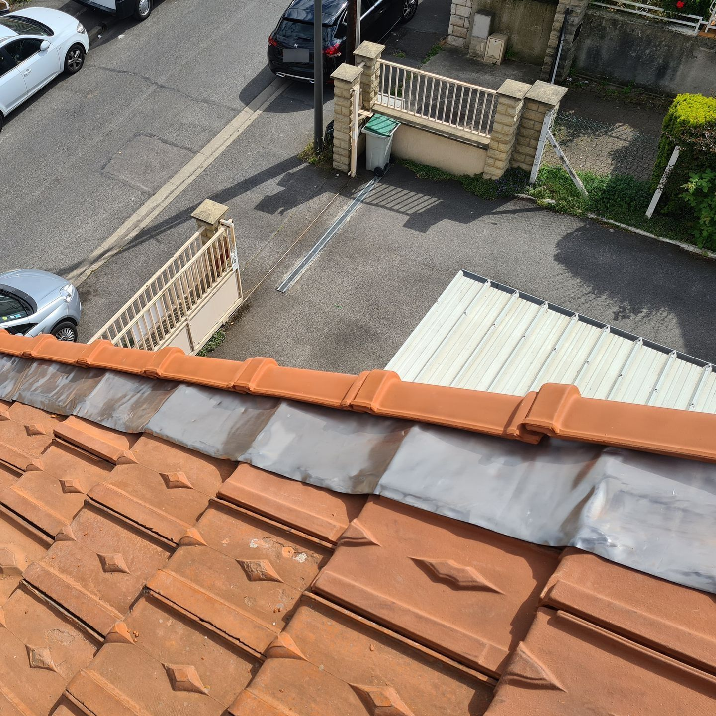 Vue depuis les toits d'une rue résidentielle, avec des tuiles en terre cuite au premier plan et des voitures stationnées en contrebas.