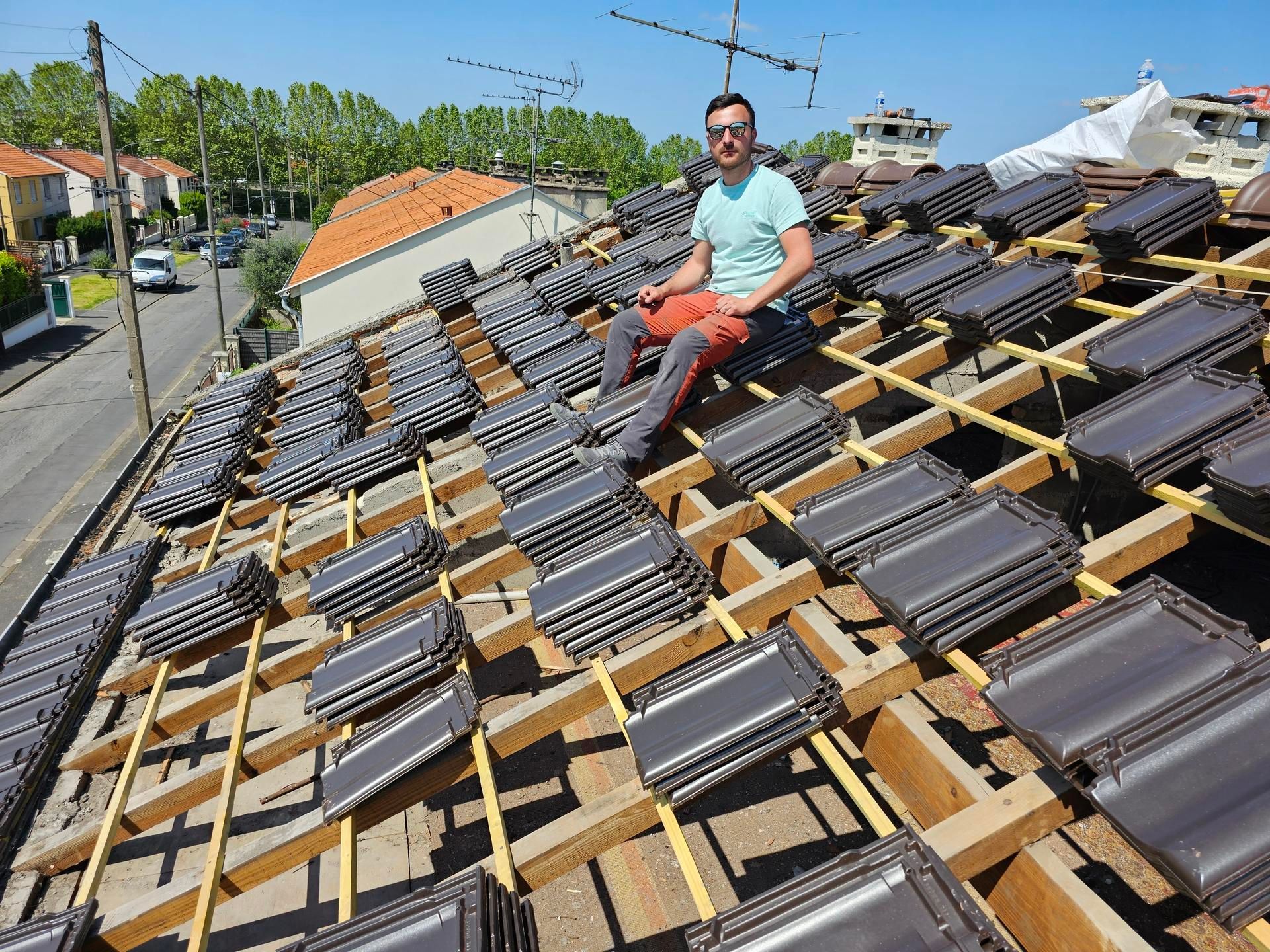 Ouvrier assis sur un toit en pente parmi des piles de tuiles.