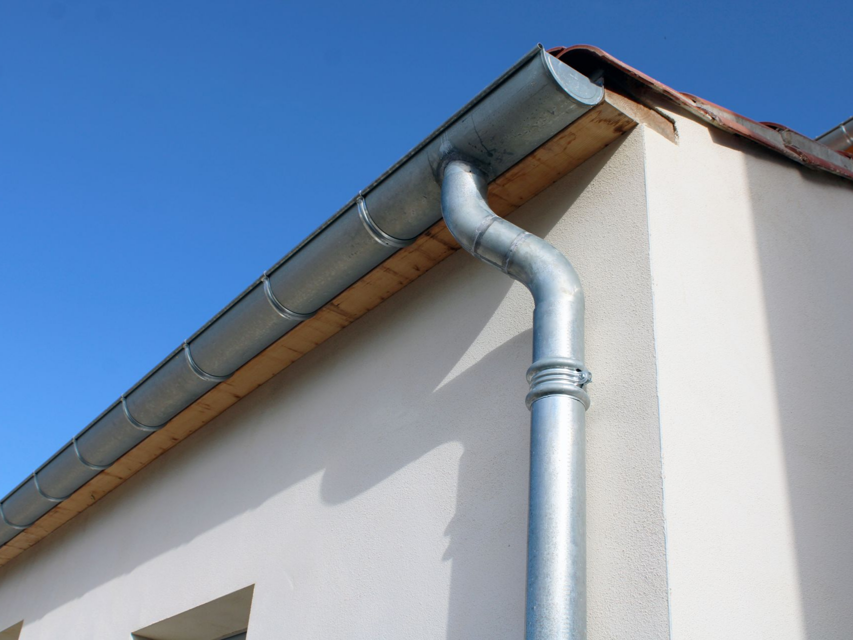 Coin d'une maison blanche avec gouttières et descentes pluviales en aluminium sur fond de ciel bleu.