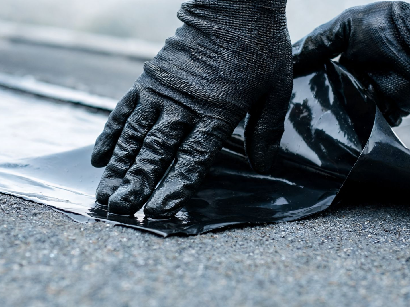 Des mains gantées appliquent une membrane de toiture noire.