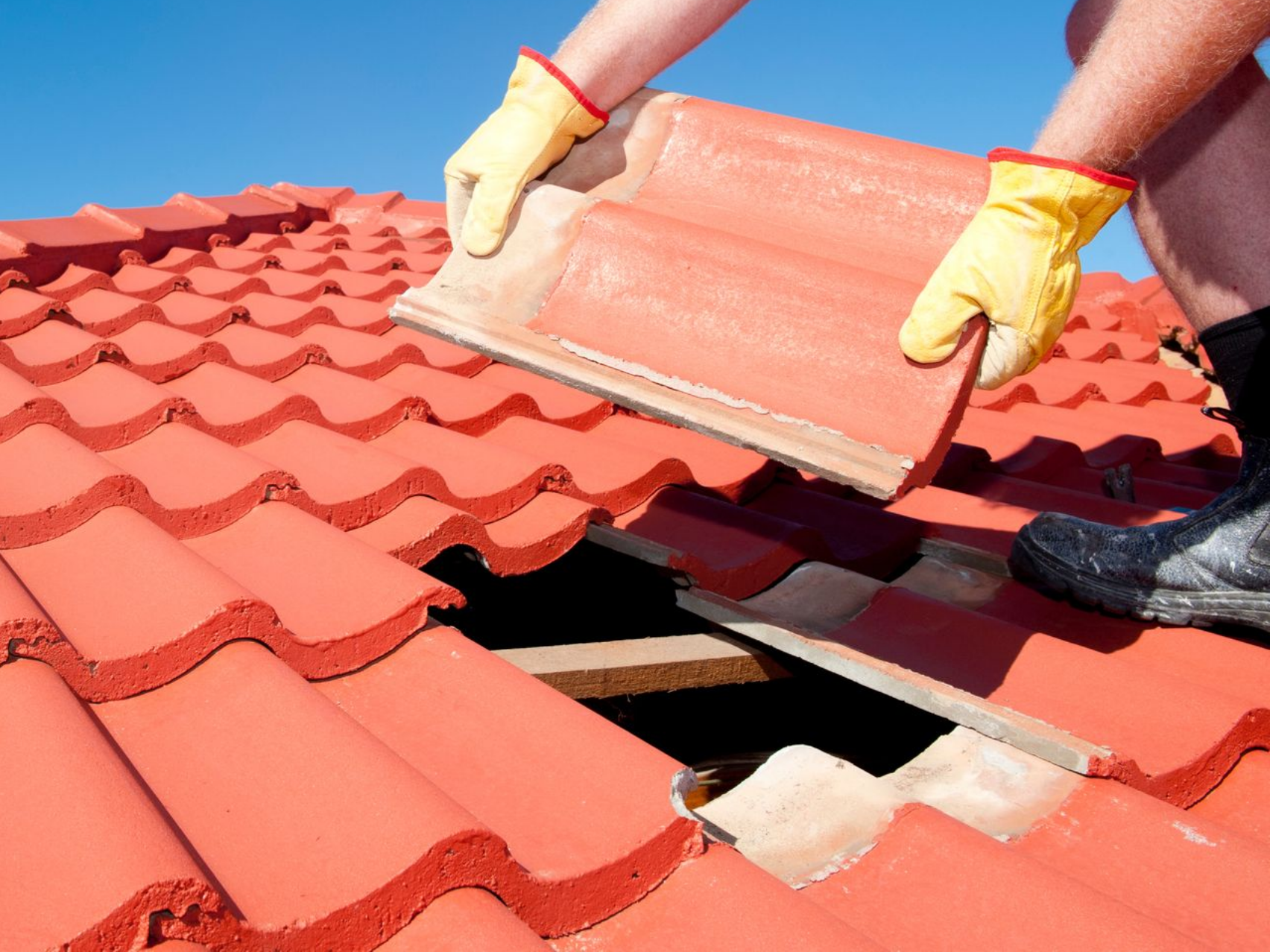 Des mains posent des tuiles rouges sur le toit d'une maison sous un ciel bleu.