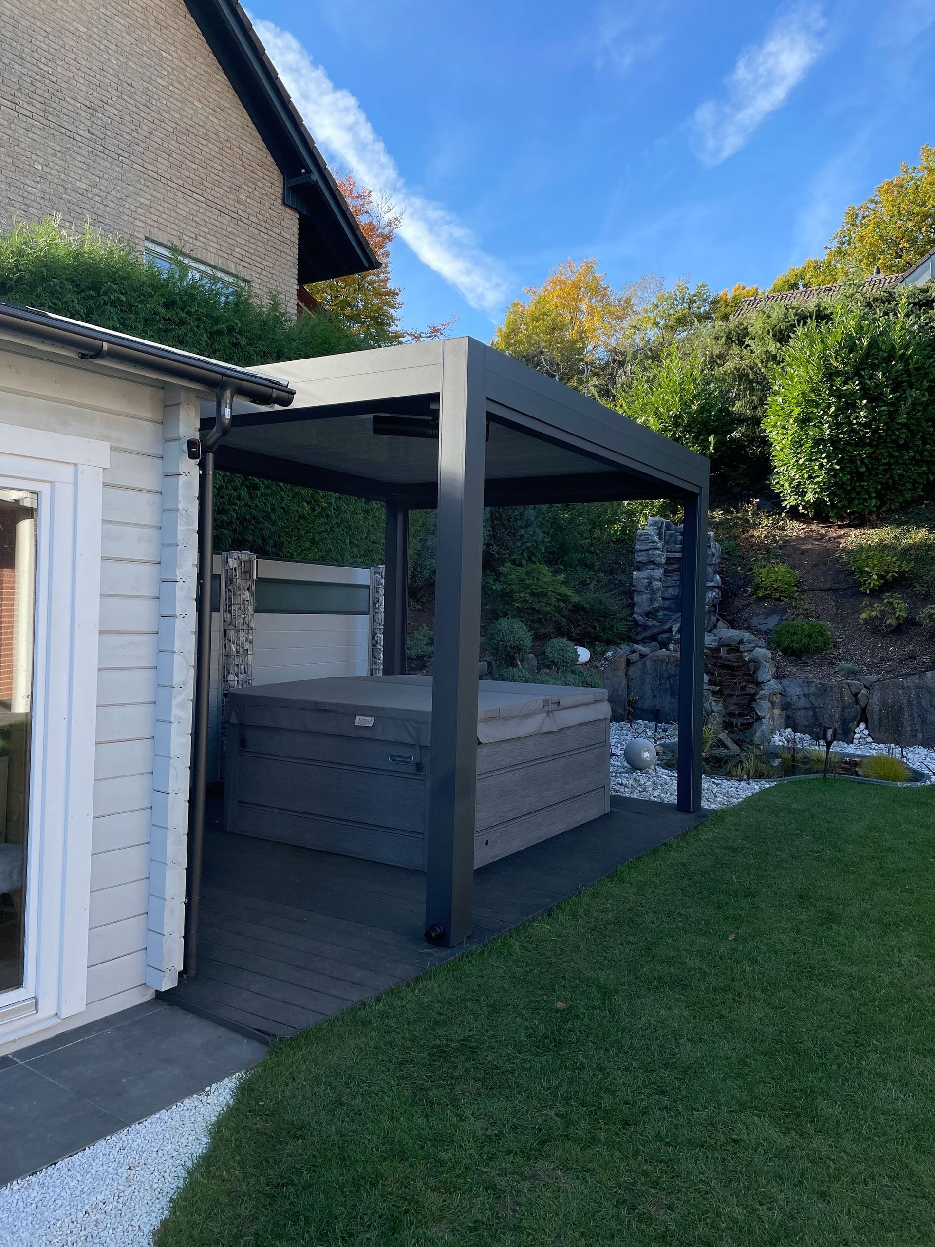 Ein Whirlpool steht unter einer grauen Pergola auf einer Holzterrasse neben einem kleinen weißen Gebäude, umgeben von grünem Gras und Bäumen.