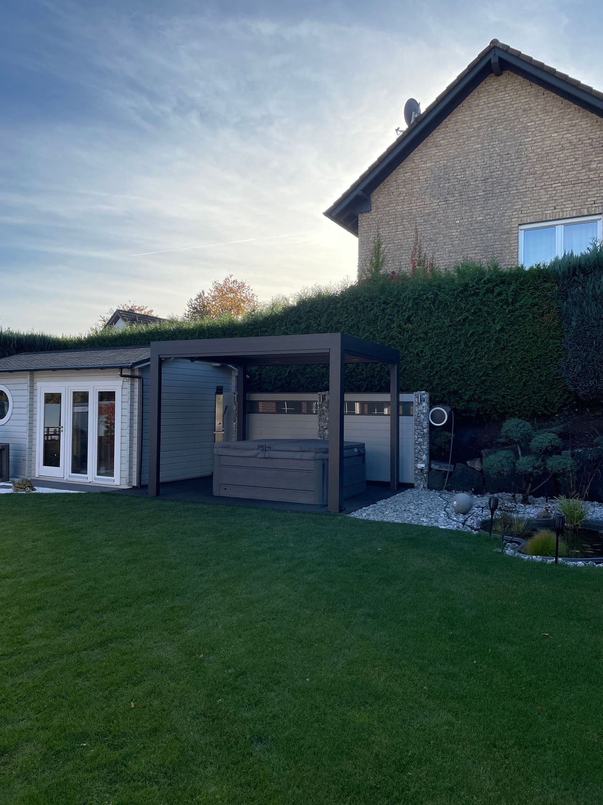 Hinterhofszene mit Whirlpool unter einer Pergola, neben einem kleinen weißen Schuppen und einem Haus mit dunklem Dach.