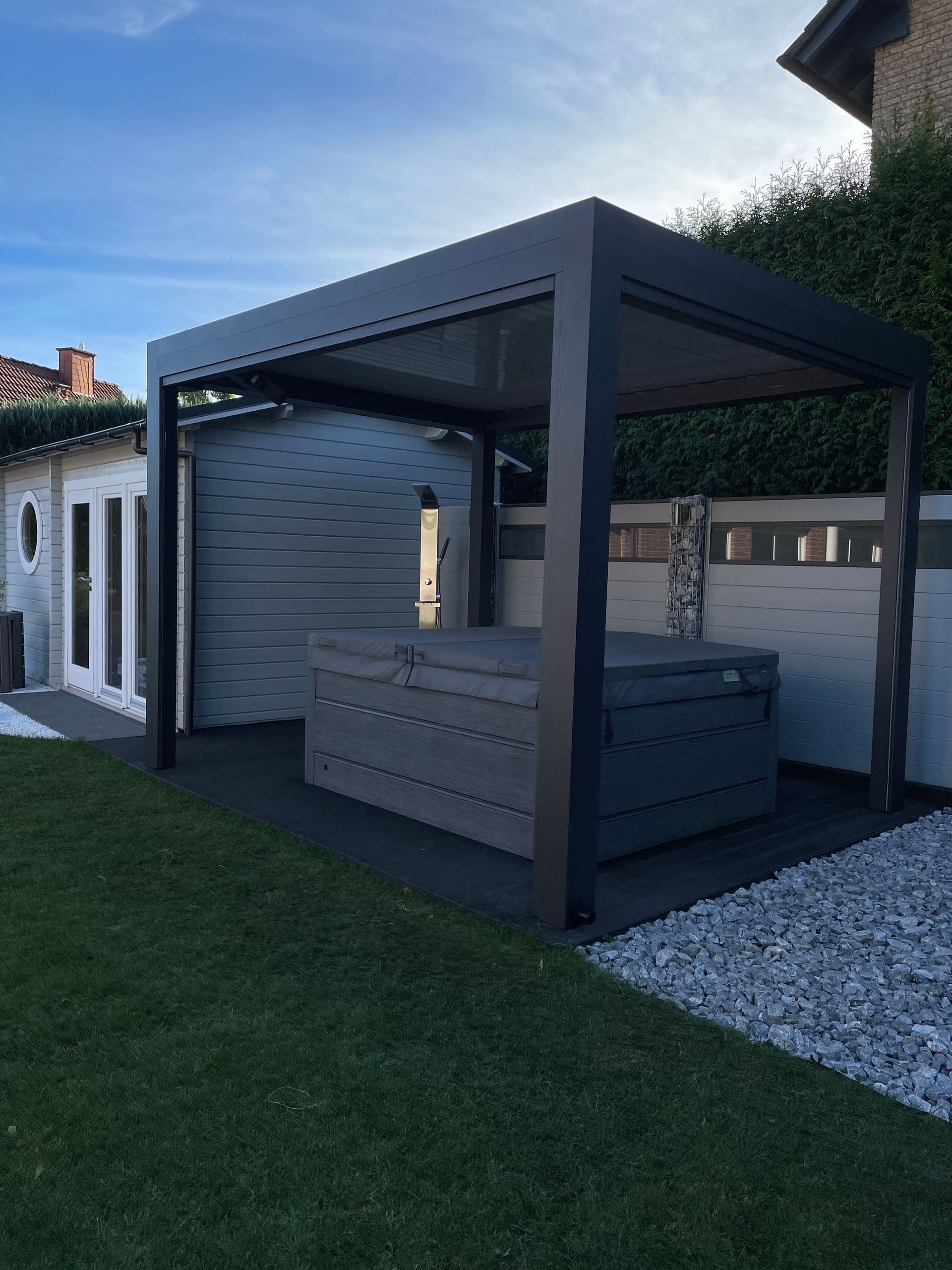Whirlpool unter einer dunkelgrauen Pergola auf einer Steinterrasse, im Hintergrund ein Schuppen und ein Zaun.