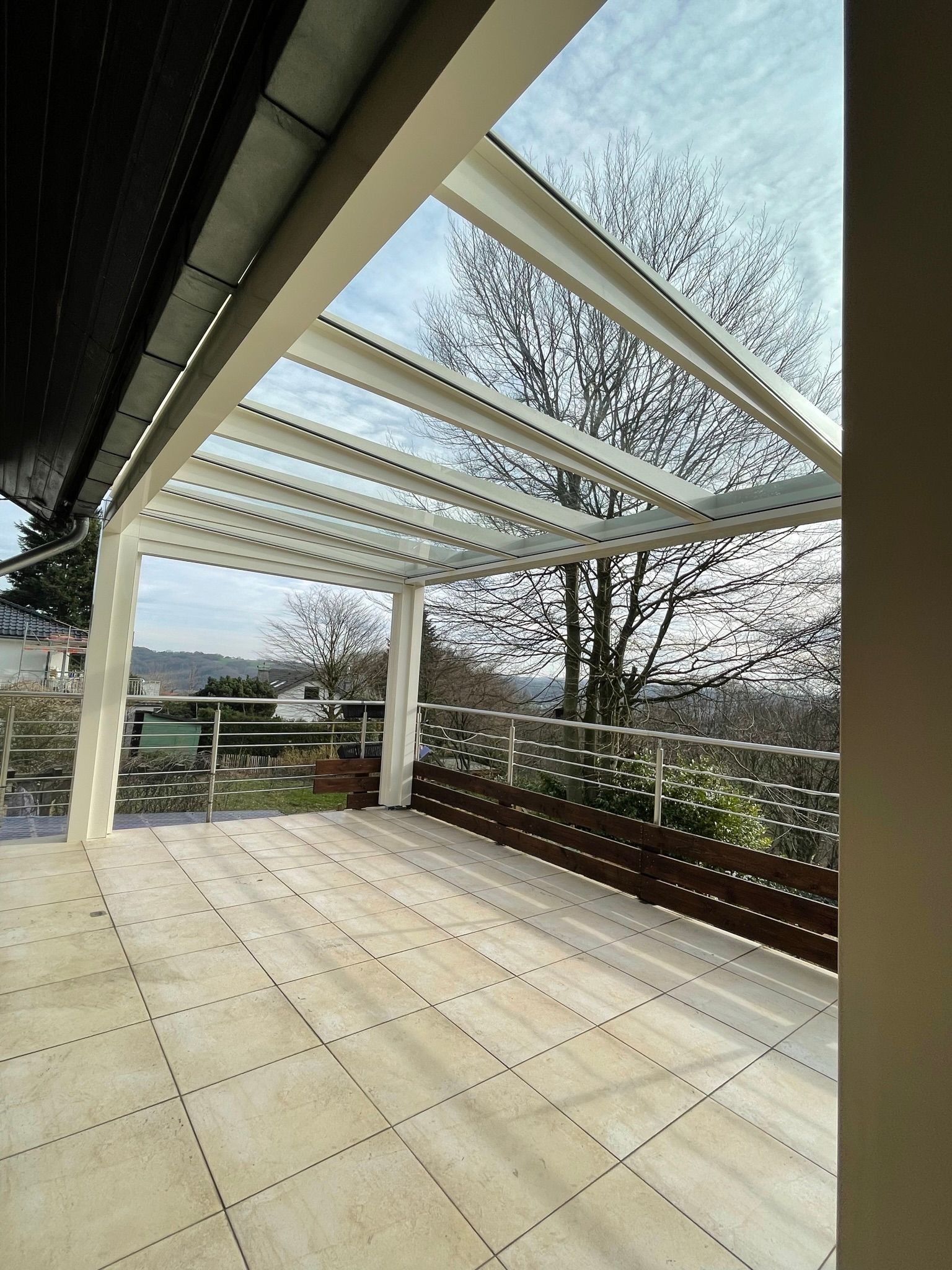 Überdachte Terrasse mit Glasdach, weißem Rahmen, braunem Geländer, beigefarbenem Fliesenboden, Blick auf Bäume und Fernsicht.