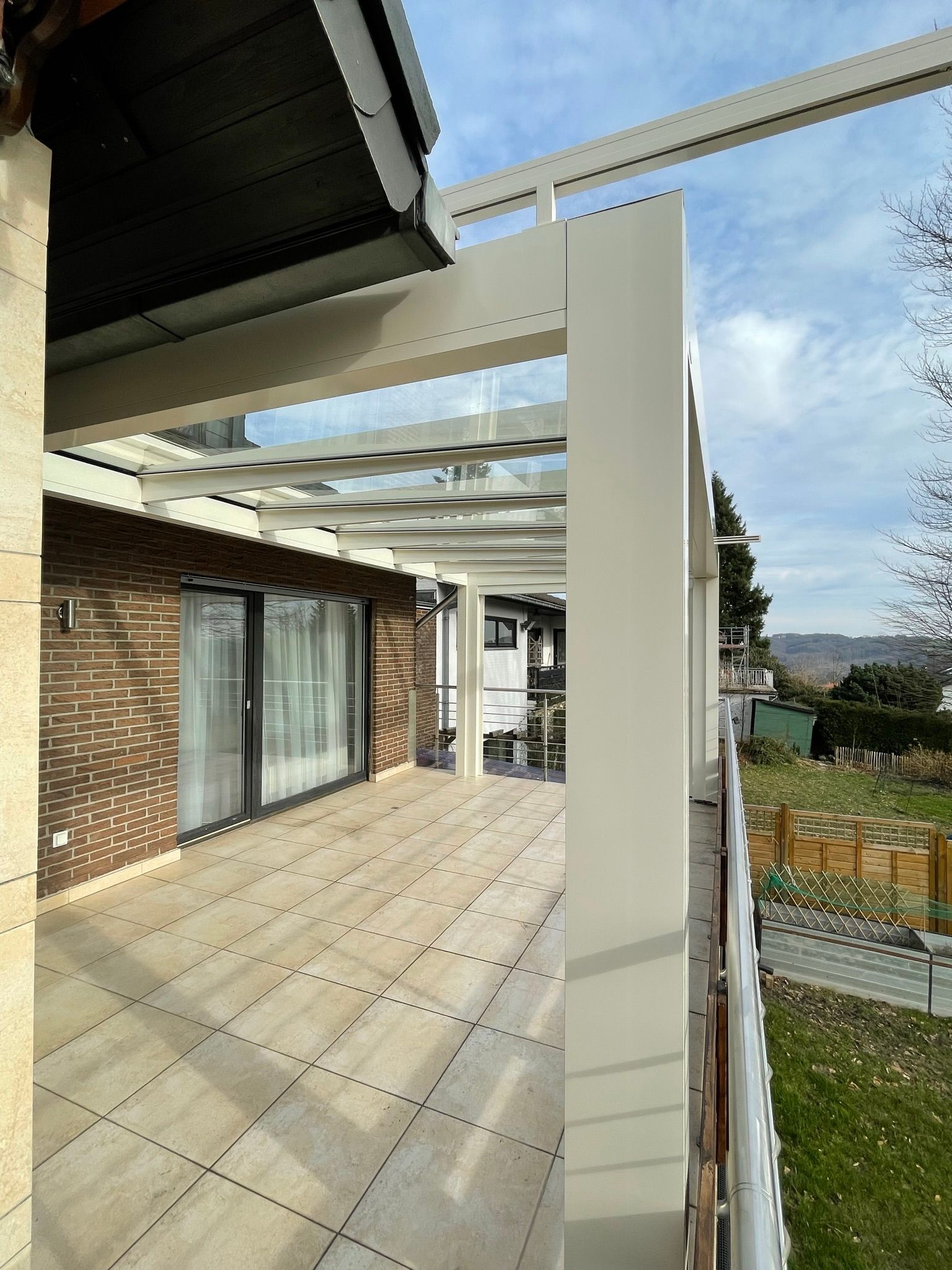 Terrasse mit Glasdach, hellen Fliesen und weißen Stützen. Blick auf ein Haus und die umliegende Landschaft.