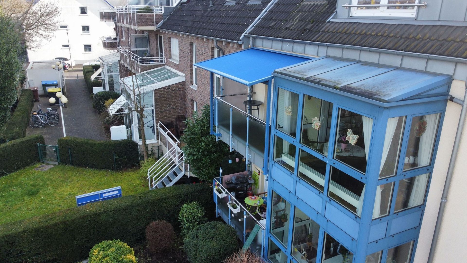 Blauer Wintergarten und Markise an einem mehrstöckigen Gebäude mit Blick auf einen grünen Rasen und die Straße.