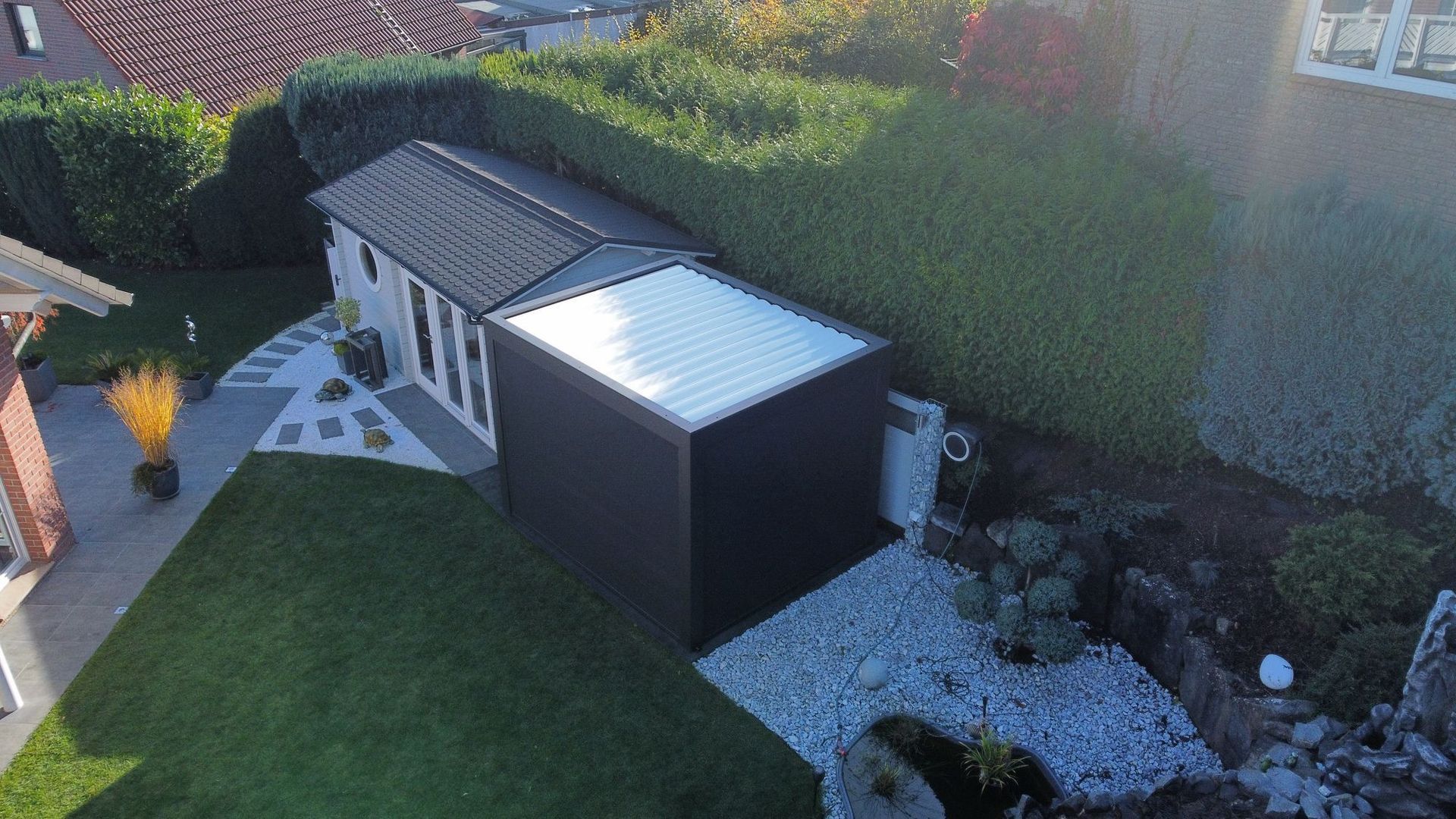 Ein modernes, dunkelfarbiges Gartengebäude mit Glasdach, neben grünem Rasen und Büschen.