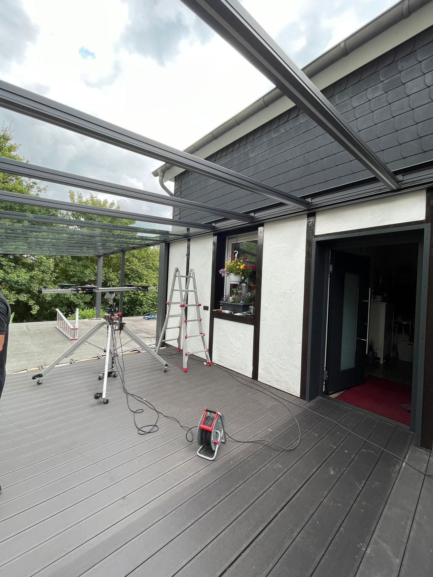 Außenterrasse mit Pergola, an der gearbeitet wird; Leiter, Geräte und Haus mit schwarzer Verkleidung.