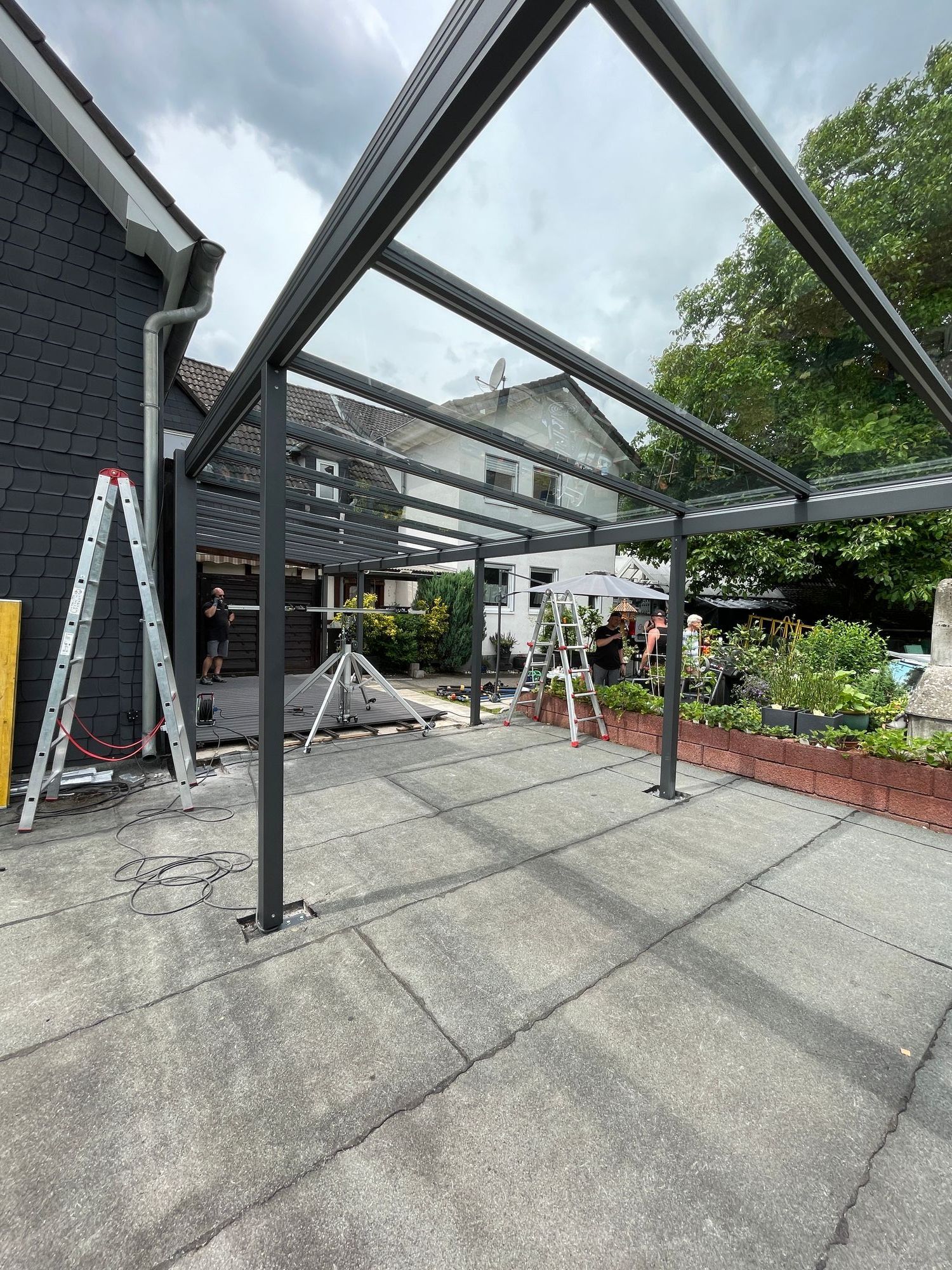 Auf einer Betonplatte neben einem dunkelgrauen Haus wird eine graue Pergola aufgebaut.
