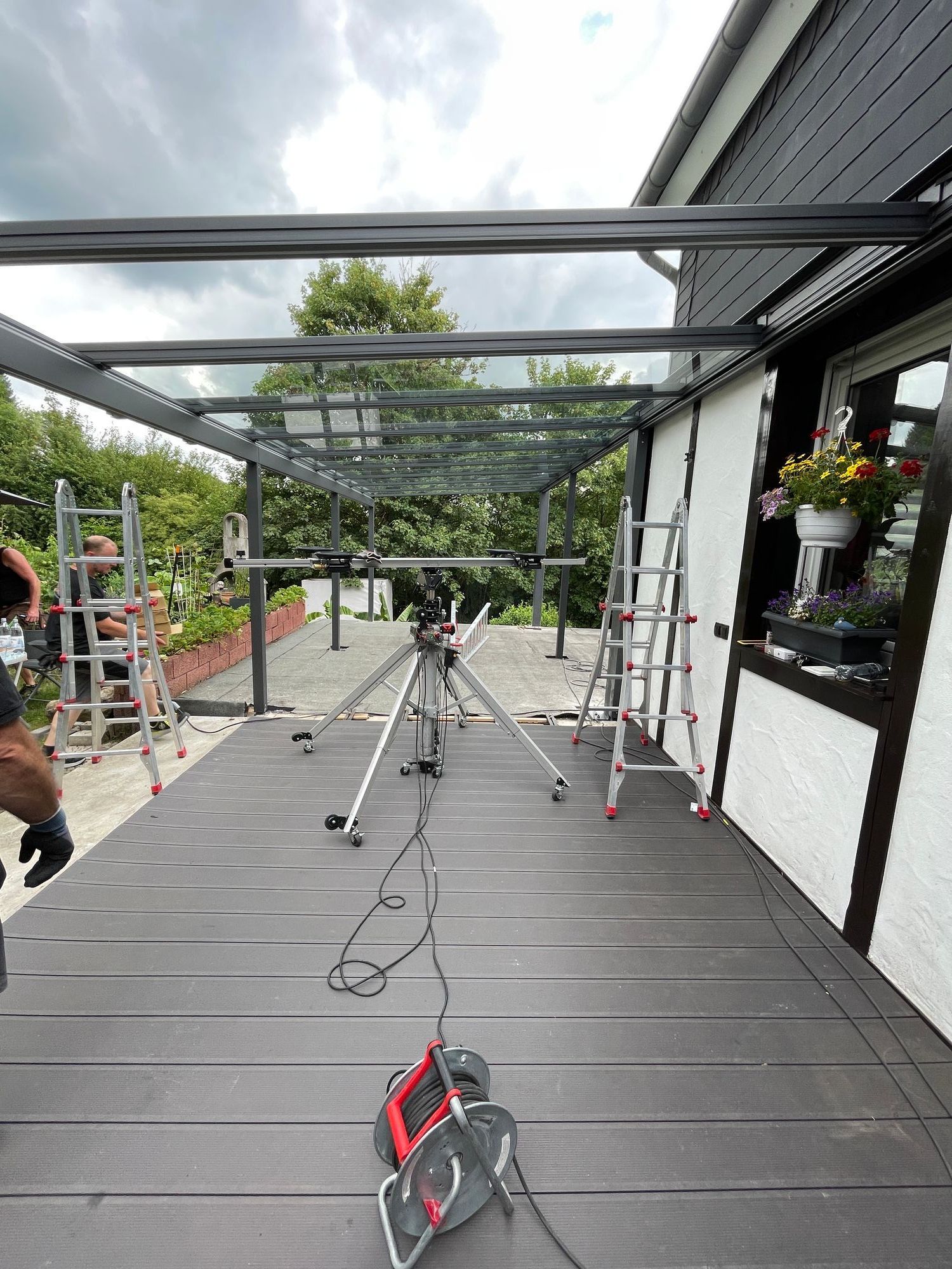 Terrasse mit Glasdach; Leitern, Werkzeug und ein Stativ sind vorhanden. Bewölkter Himmel.