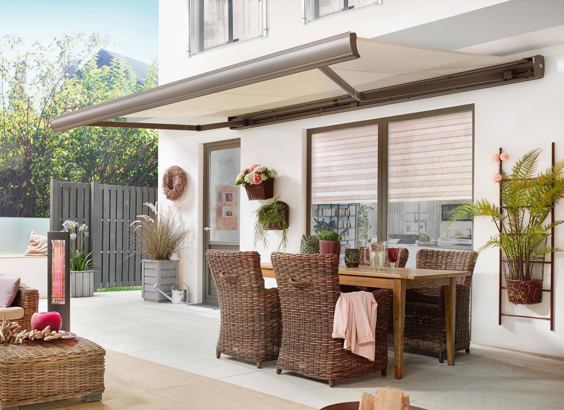 Terrasse mit ausfahrbarer Markise, Esstisch und Korbstühlen. Pflanzen und Heizstrahler schmücken den Bereich.