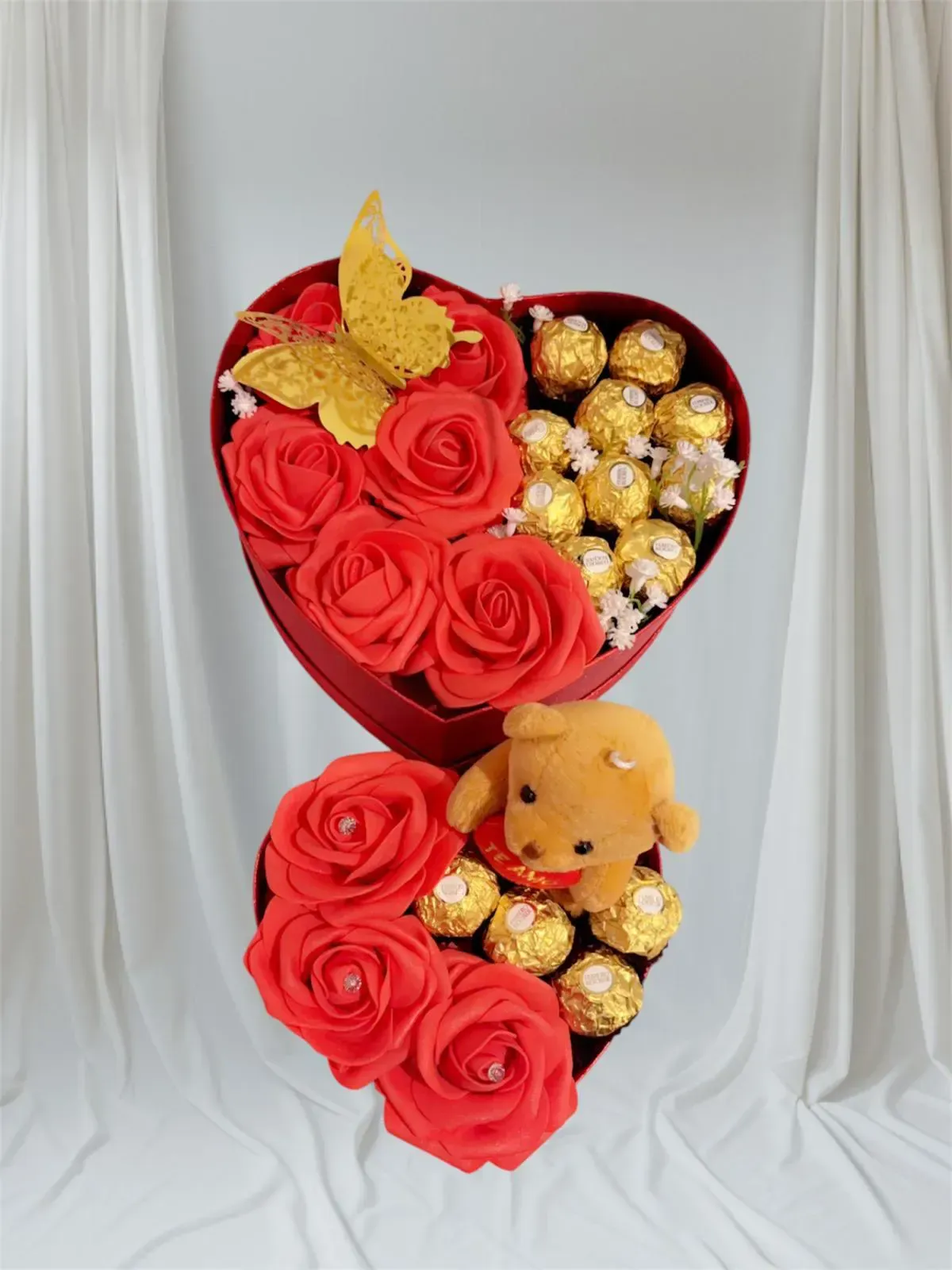 Cajas en forma de corazón llenas de rosas rojas, chocolates, un osito de peluche y una mariposa dorada como decoración.