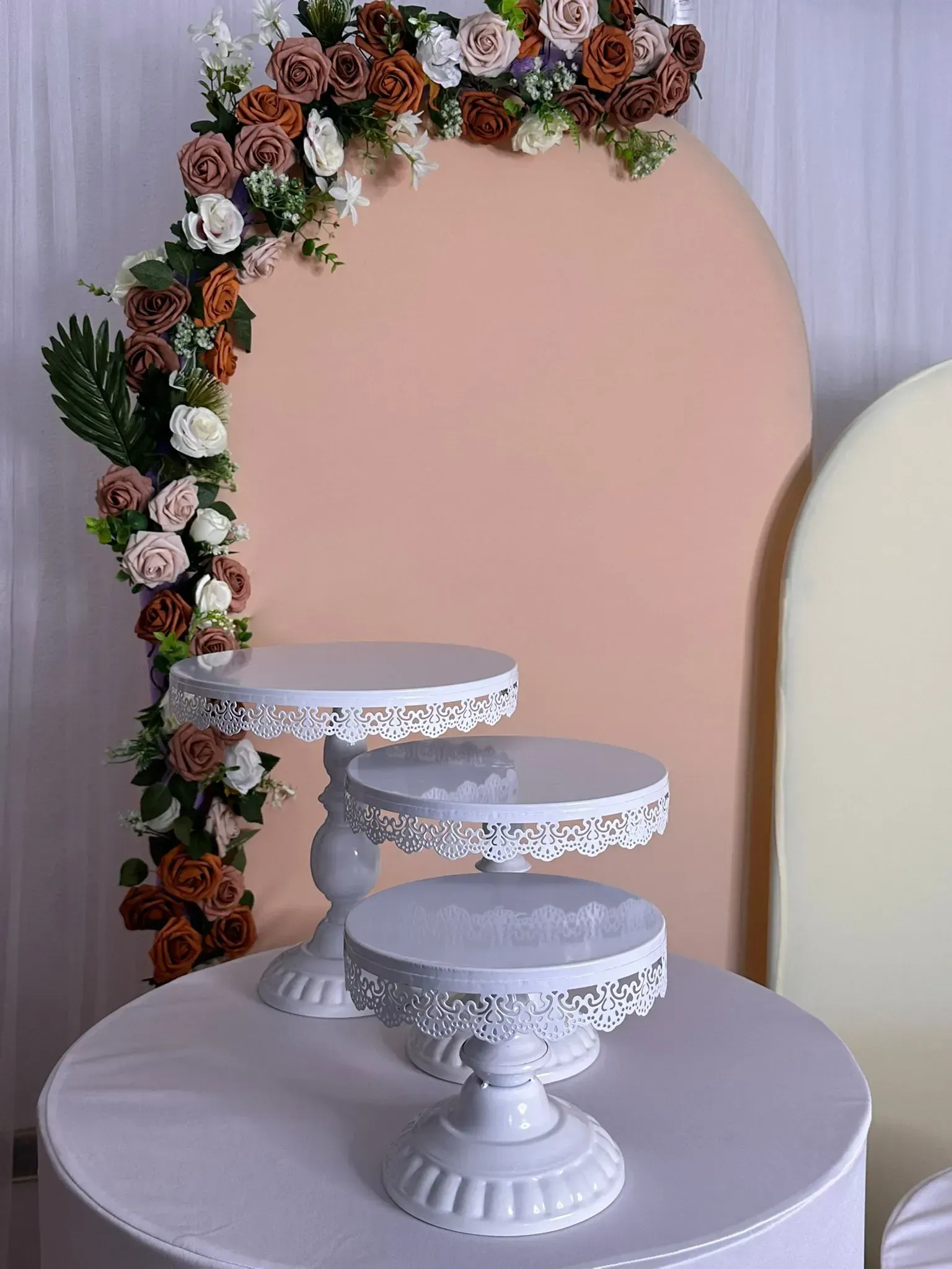Soportes para pasteles blancos de varios niveles sobre una mesa redonda, con un arco floral como fondo.