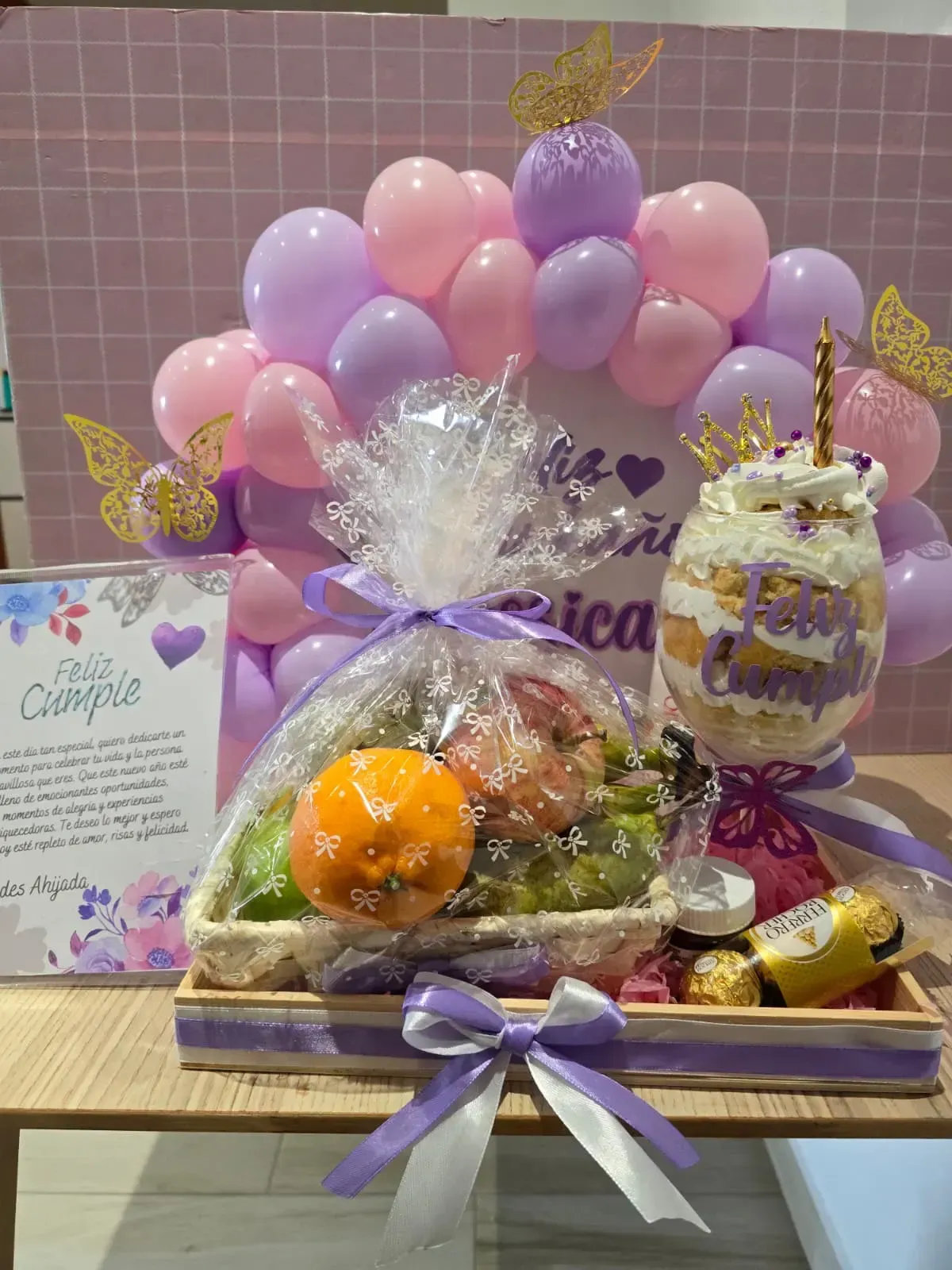Cesta de regalo con fruta, postre, globos y tarjeta en tonos morados y rosas.
