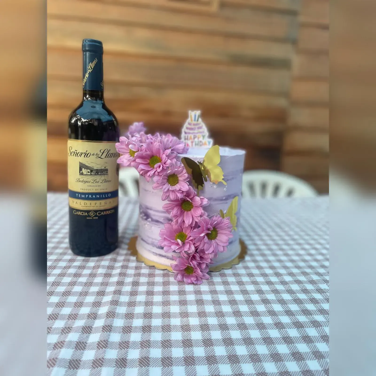 Pastel decorado con flores rosas y una botella de vino sobre un mantel a cuadros.