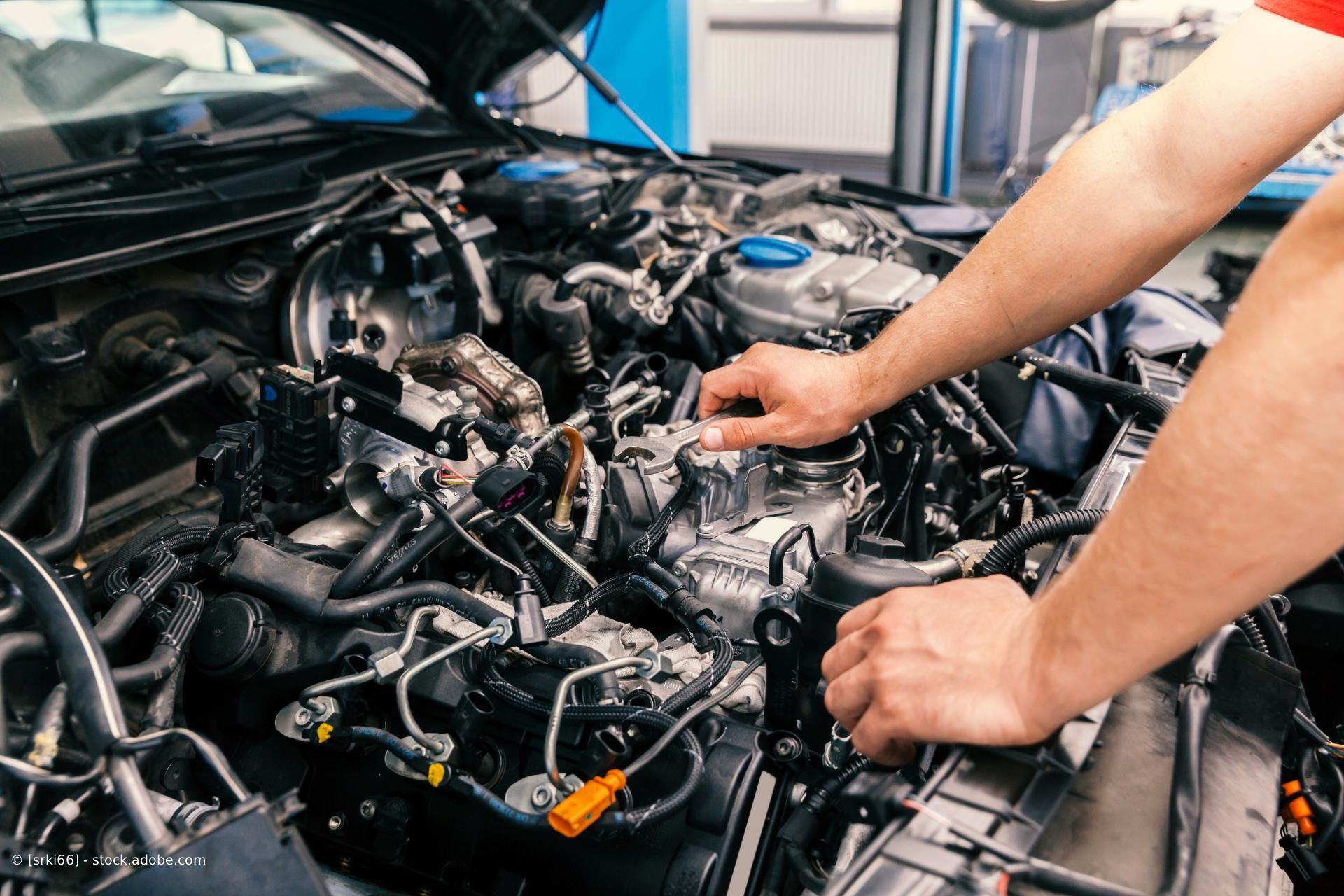 Ein Mechaniker arbeitet in einer Werkstatt unter der Motorhaube eines Autos und berührt dabei freiliegende Motorkomponenten.