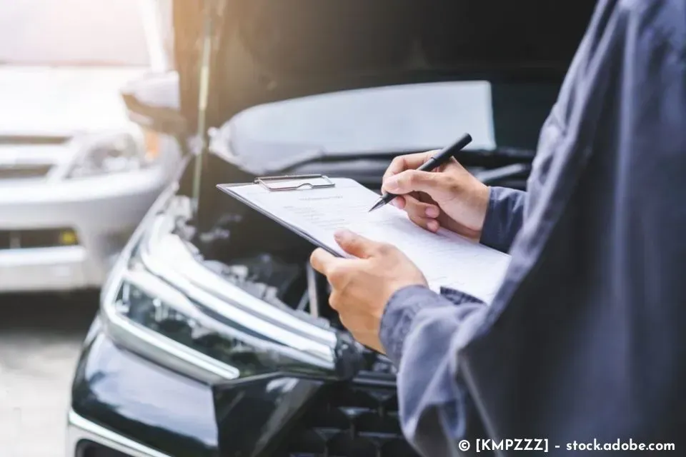 Eine Person untersucht in einer Garage einen schwarzen Wagen und schreibt dabei auf einem Klemmbrett.