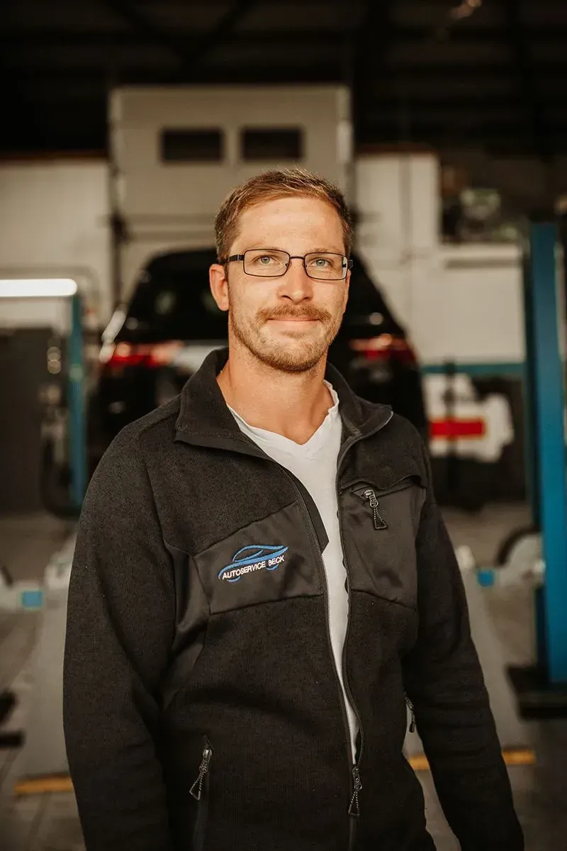 Ein Mann mit Brille und schwarzer Jacke steht in einer Garagenwerkstatt; im Hintergrund ist ein Fahrzeug zu sehen.