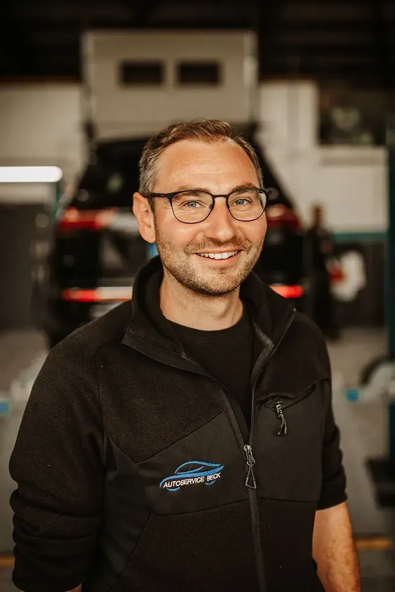 Ein lächelnder Mann mit Brille und schwarzer Jacke steht in einer Werkstattgarage.