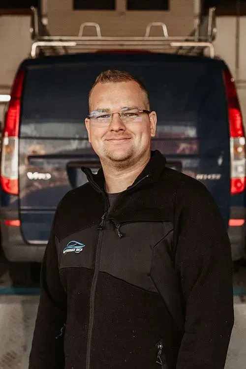 Ein Mann in einer schwarzen Jacke steht vor einem dunklen Lieferwagen und lächelt in die Kamera.