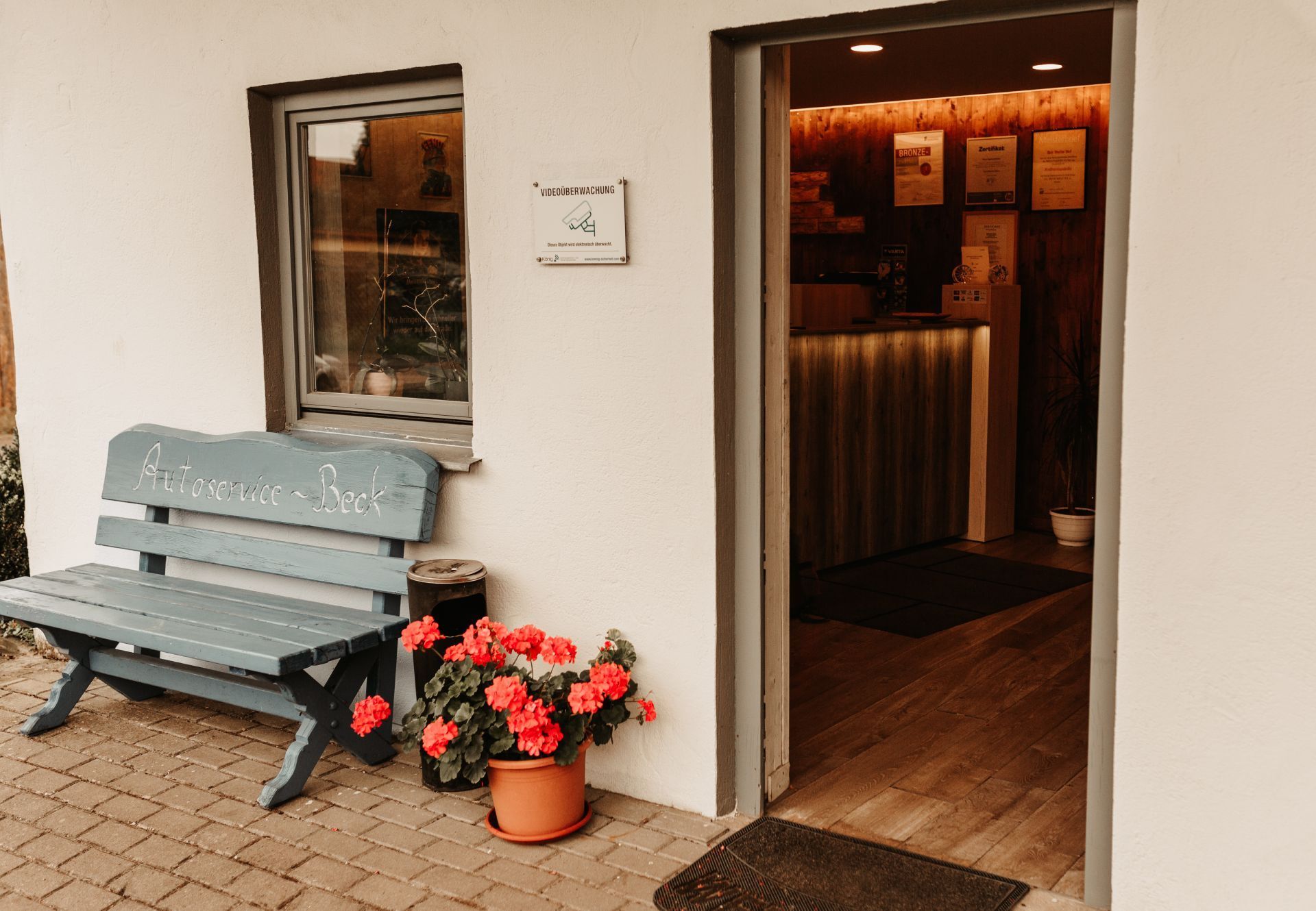 Kleiner Café-Eingang mit grauer Bank, Topfblumen und warmer Innenbeleuchtung, die durch die offene Tür sichtbar ist