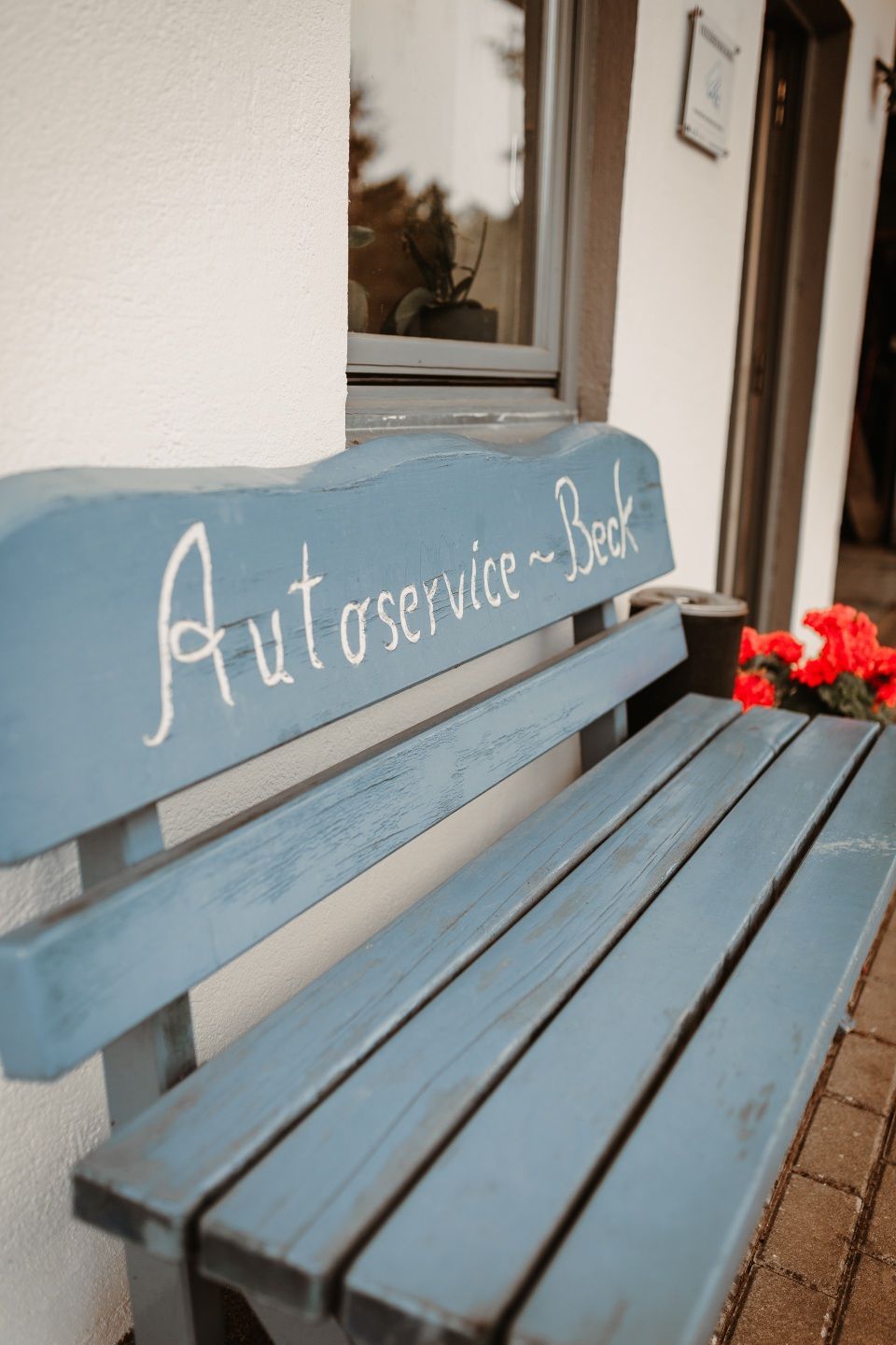 Blaue Holzbank mit der Aufschrift „Autoservice“ auf der Rückenlehne neben einer Wand und roten Blumen