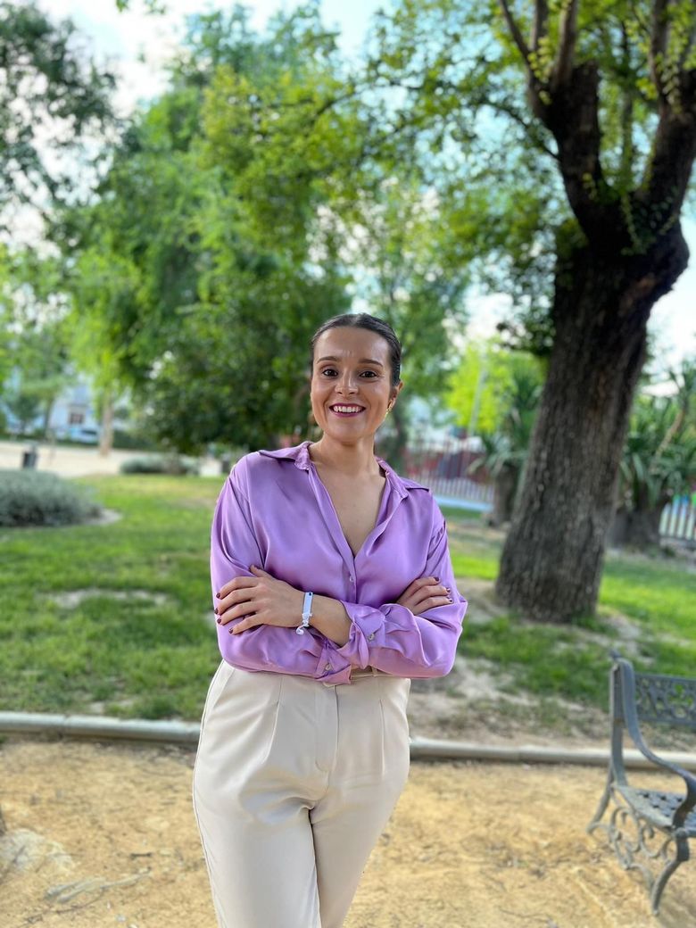 Mujer con camisa lavanda y pantalones beige sonríe, con los brazos cruzados, en un parque con árboles.