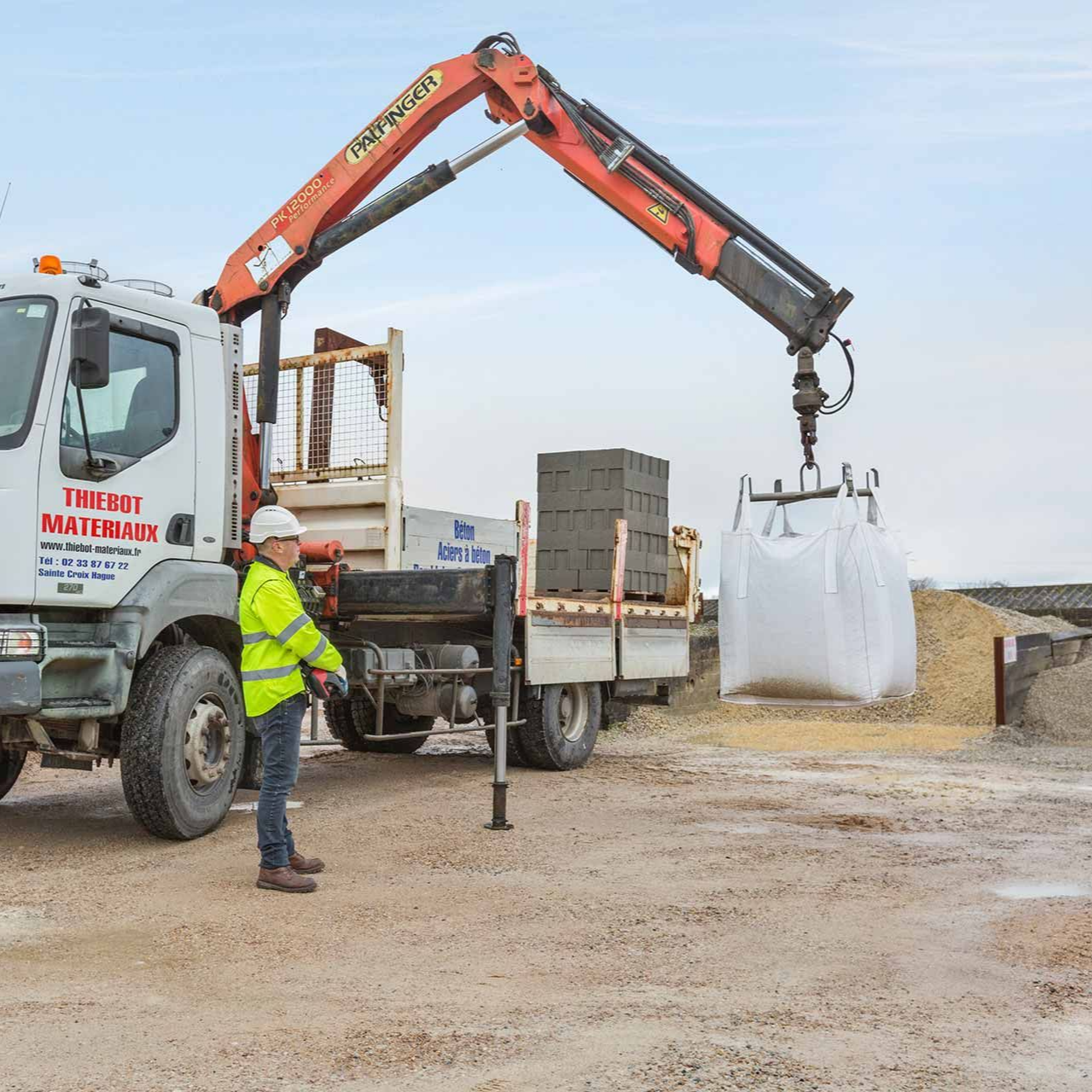 Un ouvrier utilise un camion-grue pour soulever un grand sac de matériaux sur un chantier de construction.