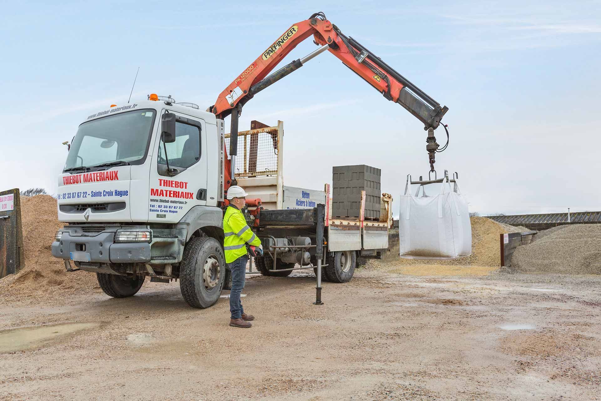Un camion équipé d'une grue soulève un grand sac blanc ; un ouvrier portant un gilet de sécurité observe sur un chantier de construction.