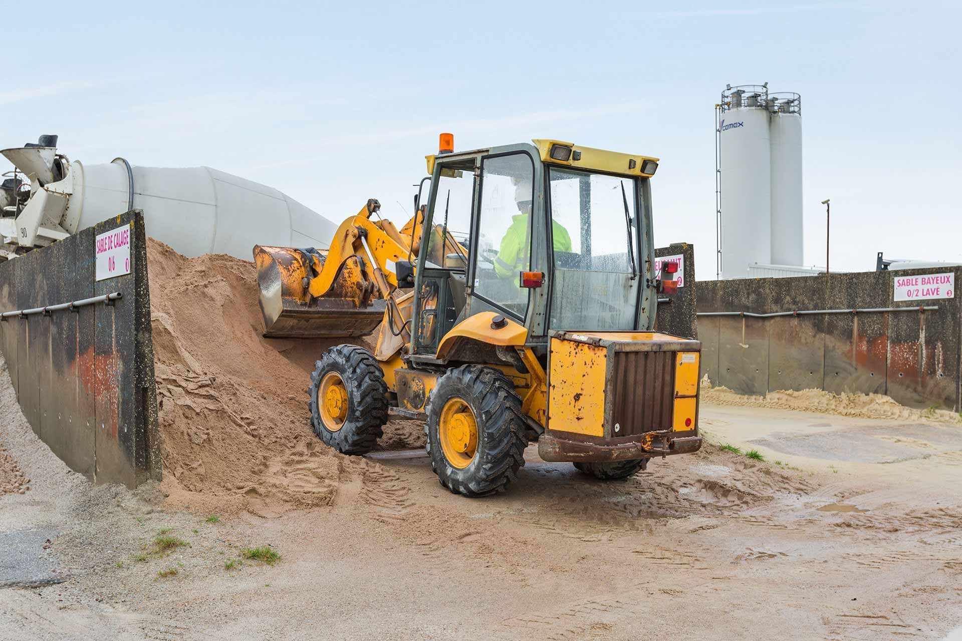 Pelleteuse jaune chargeant du sable sur un chantier de construction avec des silos en arrière-plan.