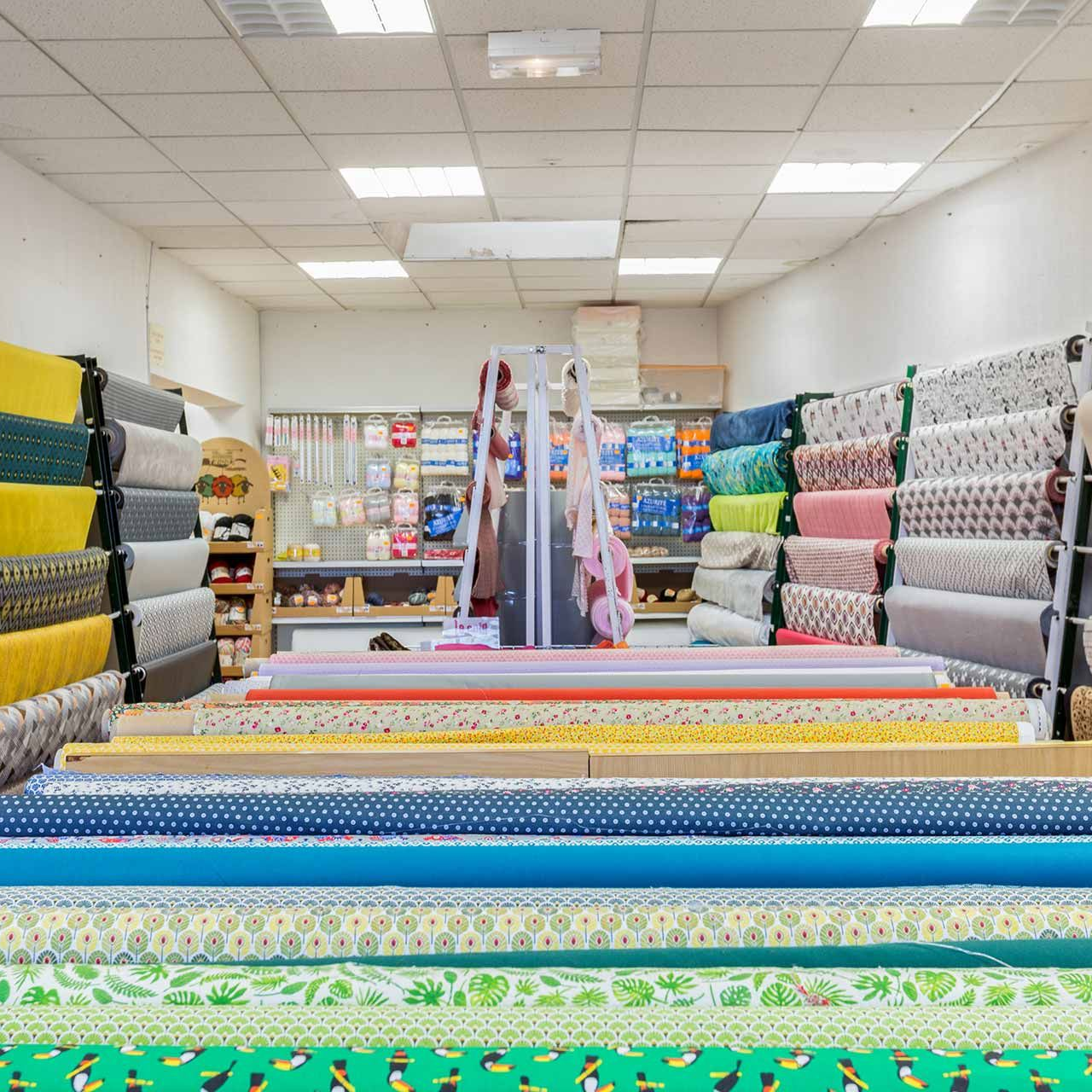 L'intérieur d'un magasin de tissus est rempli de rouleaux de tissus colorés. Des étagères en arrière-plan abritent des fournitures de couture.