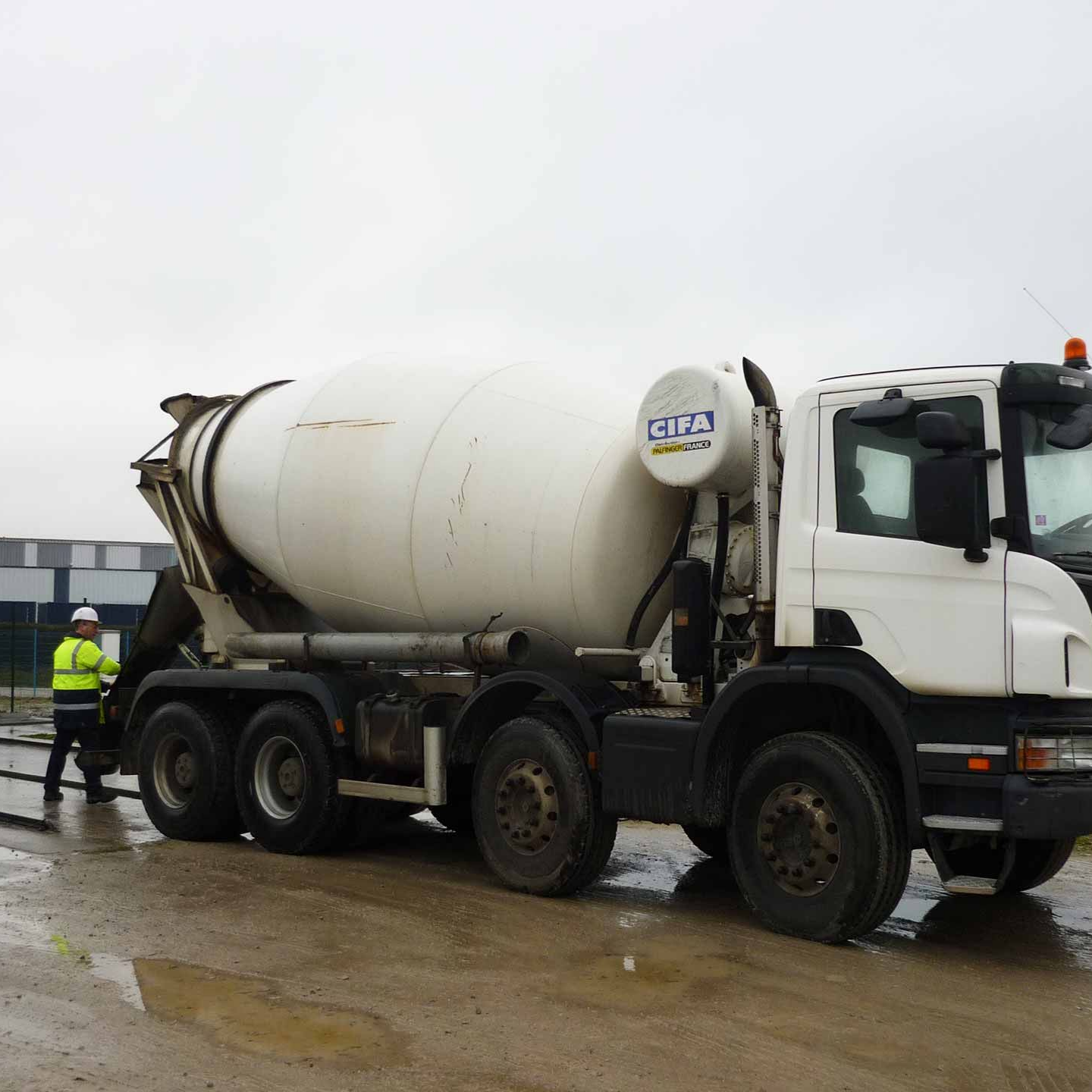 Camion de ciment sur un chantier de construction, ouvrier portant un gilet de sécurité à proximité.