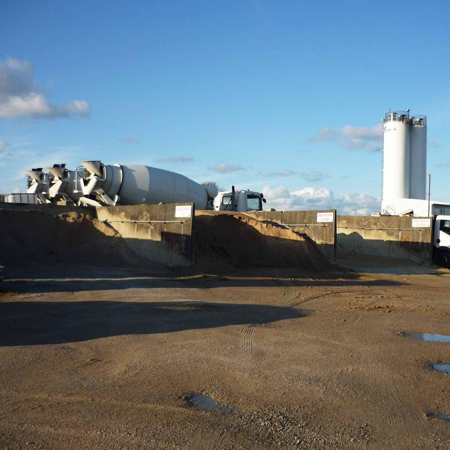 Centrale à béton : camion, silo, bétonnière, tas de graviers et un ciel bleu clair.