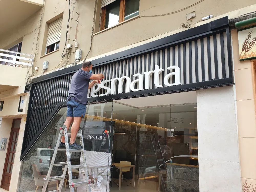 Hombre en una escalera instalando un letrero de tienda que dice "esmarta". Fachada de edificio en blanco y negro.