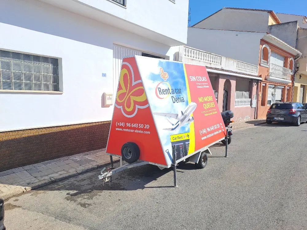Remolque rojo y blanco con anuncios de viajes estacionado en una calle cerca de edificios blancos y naranjas.