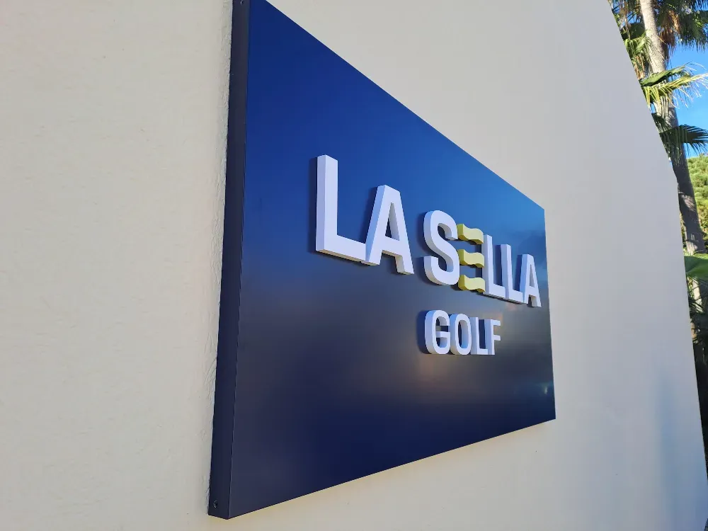 Letrero de La Sella Golf sobre fondo azul, montado en una pared color crema. Letras blancas y amarillas.