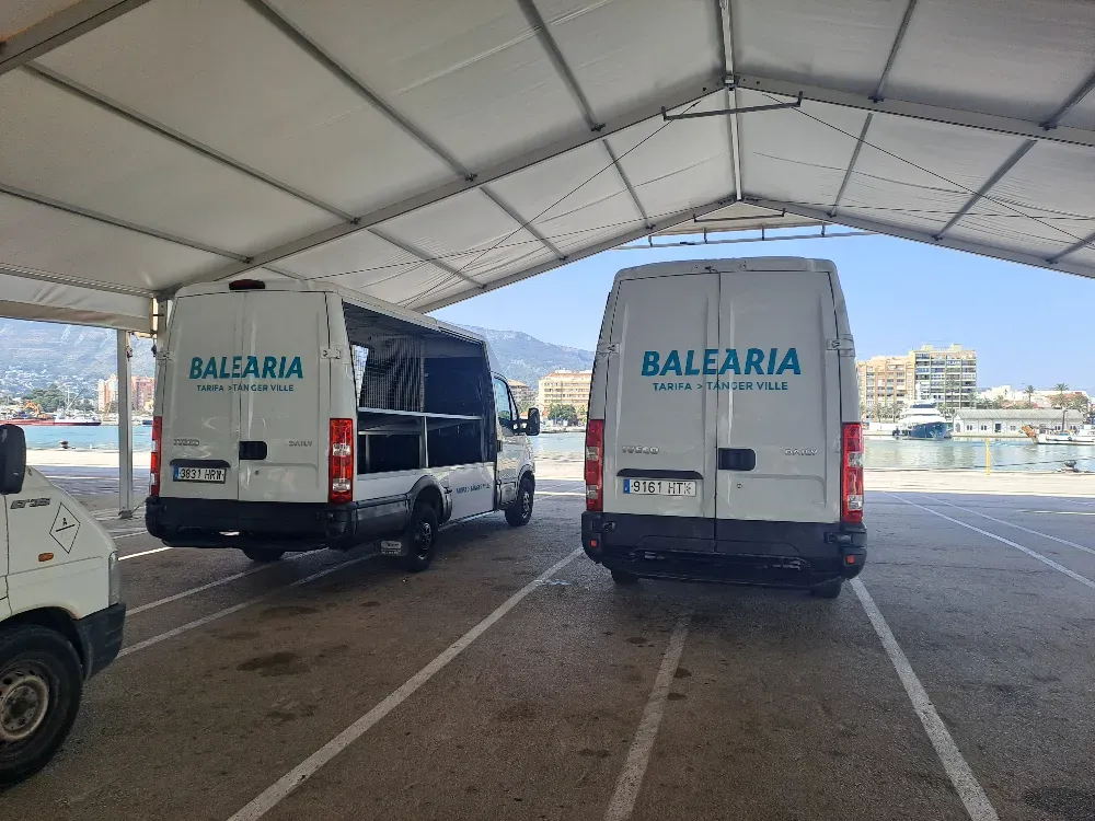 Dos furgonetas blancas de Baleària estacionadas bajo una carpa blanca. Al fondo se ve un puerto y edificios.